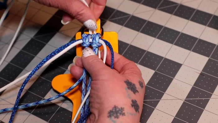 Pull the ends of the white cord down the loop, creating another cow hitch. Before tightening, add the colonial blue cord through the white cow hitch, bringing the middle to the top. Tighten everything.